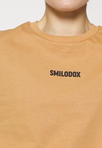 T-shirt de manga curta e decote redondo em tecido laranja claro e suave, com um logótipo preto impresso "SMILODOX" no peito.