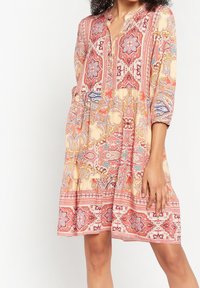 Mönstrad klänning med krage, knappar och trekvartslånga ärmar. Har en blandning av röda, rosa och gräddfärgade nyanser med invecklade mönster.