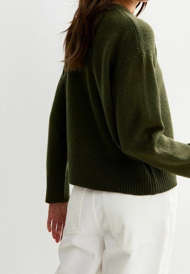 Femme portant un pull à manches longues vert foncé et un pantalon blanc, vue de dos devant un mur blanc uni.