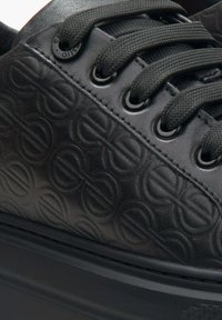 Black leather sneaker with embossed circular patterns, flat black laces, and metal eyelets. Features a thick black rubber sole.