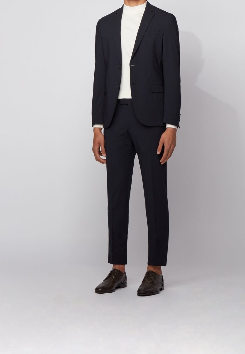 Homme portant un costume noir ajusté avec une chemise blanche à col montant et des chaussures habillées marron foncé, debout devant un fond clair uni.
