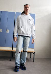 Haut à capuche gris avec une poche frontale et imprimé "1894", associé à un jean bleu clair déchiré et des baskets noires avec des accents bleus.