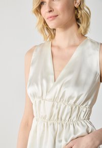 Robe en mélange de soie dans un ivoire doux, avec un décolleté en V profond, une taille froncée et une texture lisse. Soulignée par une coupe décontractée et un design fluide.