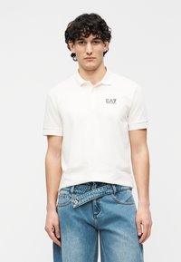 Polo branca de manga curta com gola, apresentando um pequeno logótipo EA7 no peito. Usada com jeans de ganga azul.