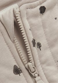 Un tissu beige clair présente une fermeture éclair avec une fermeture en plastique blanche. Des motifs d'arbres noirs et de cœurs sont imprimés sur le matériau.