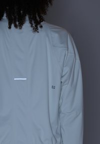 Personne portant une veste gris clair avec "S2" imprimé sur le côté droit du dos et des cheveux bouclés partiellement visibles.