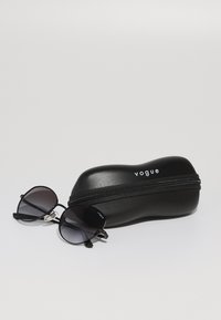 Óculos de sol pretos com lentes degradê e armações redondas ao lado de um estojo preto com fecho zip, featuring o logótipo "Vogue" em branco.