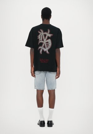 Svart oversized t-skjorte med teksturert grafisk design på baksiden, kombinert med lyseblå denimshorts og hvite ankelstrømper. Svarte sko fullfører stilen.