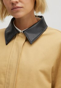 Beige Jacke aus Baumwoll-Leinwand mit einem Kragen aus schwarzem Leder. Die Textur ist glatt, mit einem Knopfverschluss und subtilen Steppdetails.