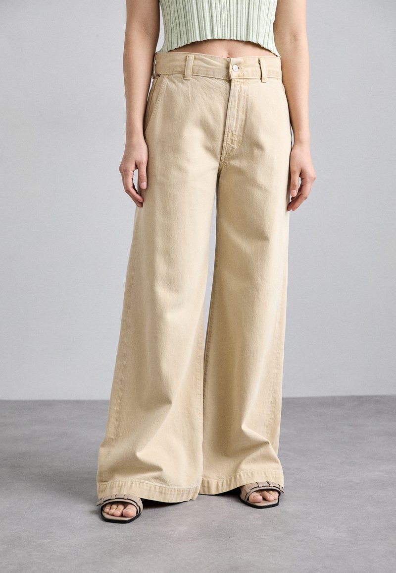 Pantalon large beige en coton doux, doté d'une taille haute, de passants de ceinture et d'une fermeture à bouton, associé à des sandales noires à lanières.