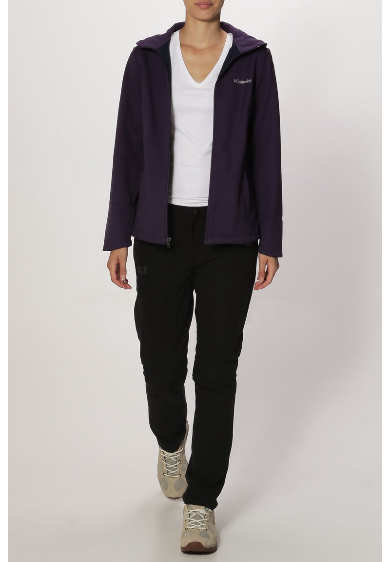 Personne portant une veste violette zippée, un t-shirt blanc col en V, un pantalon noir et des chaussures de randonnée beiges, debout devant un fond uni.