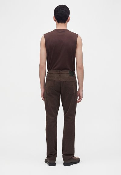 Homme debout de dos portant un haut sans manches marron foncé, un pantalon marron avec des poches à l'arrière et des chaussures marron bicolores sur un fond uni.