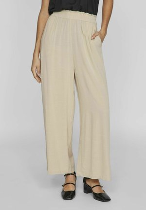 Pantalones de pierna ancha beige con cinturilla elástica, que cuentan con dos bolsillos laterales. La tela es suave y ligera. Combinados con zapatos de tacón cuadrado negros.