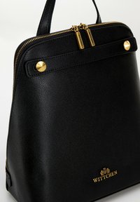 Sac à main en cuir noir avec quincaillerie dorée, présentant une forme structurée, une fermeture éclair et un logo de marque embossé en bas.