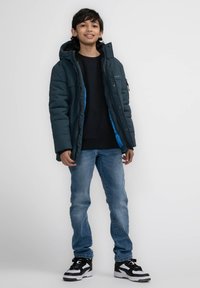 Mörkblå puffjacka med blåfoder, svart sweatshirt, ljusblå jeans och svart-vita sneakers; stående pose framför vit bakgrund.