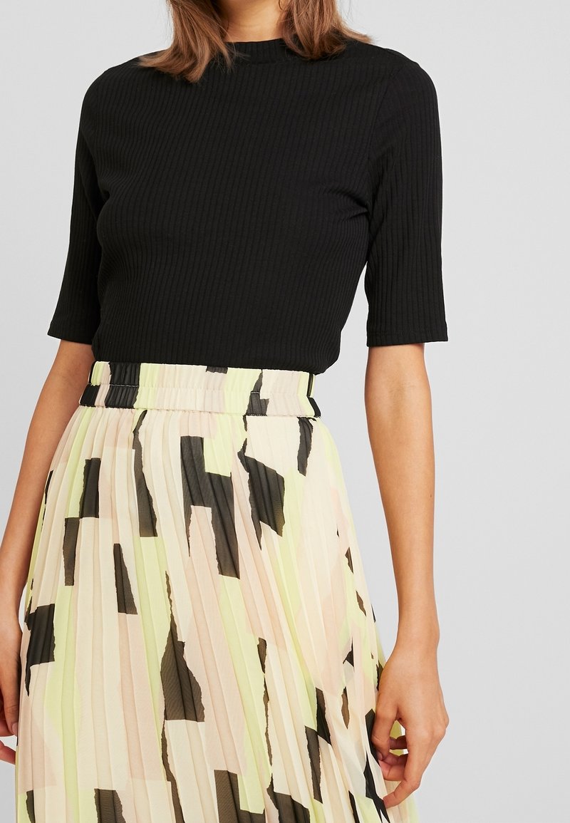 Femme portant un haut noir côtelé à manches courtes et une jupe plissée taille haute avec des motifs abstraits noirs et jaune pâle.