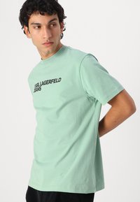Mintgrønn bomull T-skjorte med korte ermer, med en svart trykt logo "KARL LAGERFELD JEANS" over brystet. Minimalistisk design.