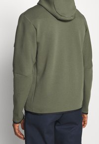 Person som bär en olivgrön huvtröja och mörkblå byxor, sedd bakifrån med händerna avslappnat vid sidorna.