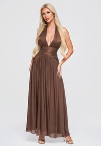 Femme blonde portant une robe marron plissée sans manches à col en V plongeant et des talons ouverts dorés, debout, une main sur la hanche.