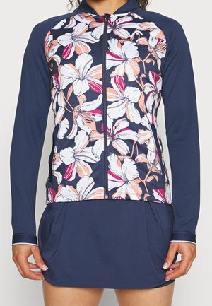 Frau trägt eine marineblaue Reißverschlussjacke mit langen Ärmeln und großem weißem sowie pfirsichfarbenem Blumenmuster auf der Vorderseite, kombiniert mit einem passenden marineblauen Rock.
