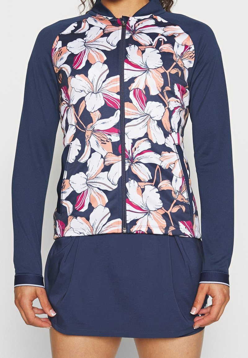 Femme portant une veste zippée bleu marine à manches longues avec un grand imprimé floral blanc et pêche sur le devant, associée à une jupe bleu marine assortie.