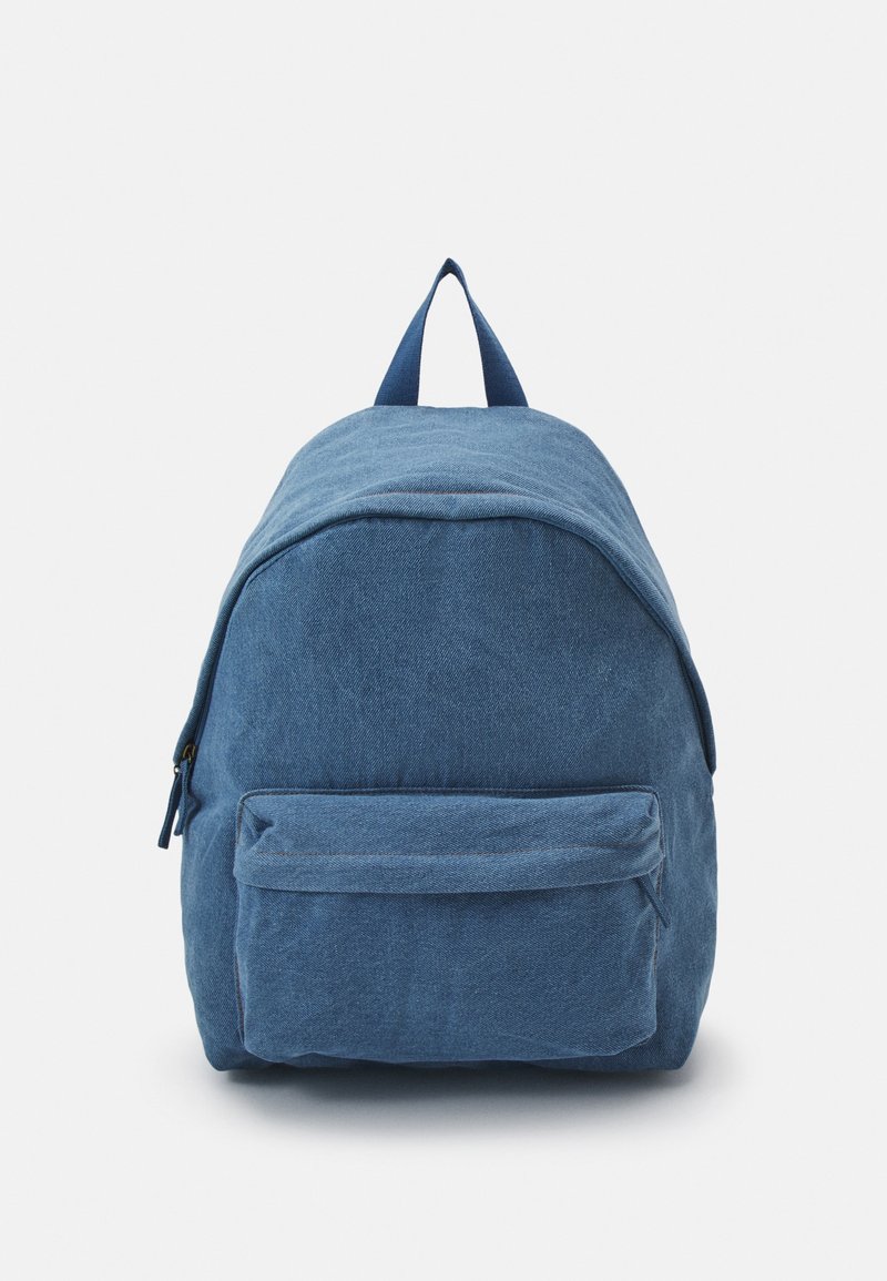 Mochila de tela azul con un bolsillo frontal con cremallera y una asa superior para llevar, mostrada de pie sobre un fondo blanco.