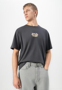 Young man wearing a dark gray "WILD SOUL" t-shirt and light gray jeans, standing with one hand in pocket, looking to the side.