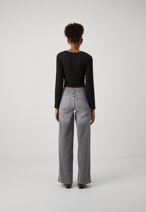 Femme debout de dos, portant un crop top noir à manches longues, un jean gris à jambes larges et des chaussures noires cloutées sur un fond uni.