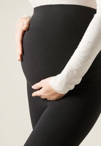 Schwarze, dehnbare Leggings umschmeicheln den Körper und werden mit einem weißen Langarmoberteil kombiniert. Der Schwerpunkt liegt auf dem runden Bauch und der glatten Stofftextur.