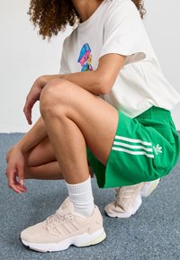 Bílý tričko s barevným potiskem, zelené kraťasy se třemi pruhy a tenisky v neutrálních odstínech. Na sobě bílé ponožky. Neformální sportovní outfit.