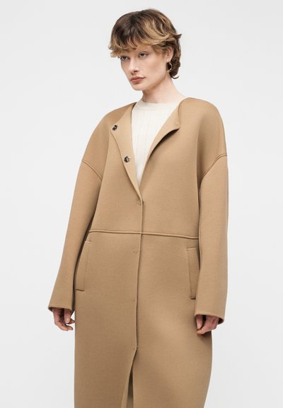 Γυναίκα με κοντά μαλλιά που φοράει μεγάλο κάμελ παλτό με μεγάλες τσέπες και μπεζ ριγωτό πουλόβερ, στέκεται μπροστά σε απλό φωτεινό φόντο.