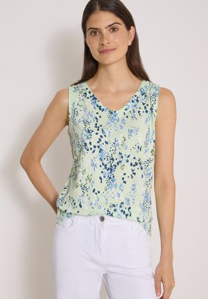 Femme aux longs cheveux bruns portant un haut sans manches vert clair avec des motifs abstraits bleus et verts et un pantalon blanc, debout avec une main sur la hanche.
