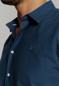 Marineblauwe button-up shirt van een gestructureerde stof, met een kraag en subtiele logodetail. Zichtbare knopen en nette stiksels.