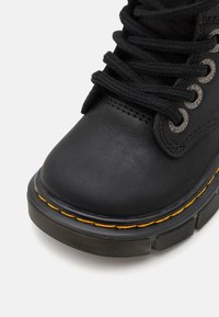 Černá kotníková kožená bota s kulatou špičkou, černými tkaničkami, kovovými průchodkami a výrazným žlutým prošíváním podél podrážky. Vrchní část s texturovaným detailem.