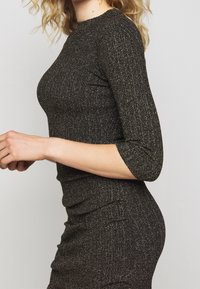 Svart ribbad klänning med metalltråd, med trekvartsärmar och en åtsittande silhuett. Slät textur och rund halsringning.