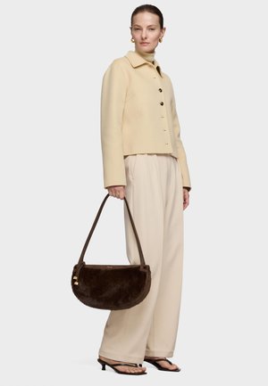 Mujer que lleva una chaqueta crema y pantalones anchos, sostiene un bolso de piel marrón y lleva sandalias negras de tacón, de pie contra un fondo liso.