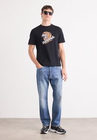 Tricou din bumbac negru cu un grafic de tigru și text, asortat cu blugi denim de culoare albastru deschis și pantofi sport negri. Modelul poartă ochelari de soare negri.