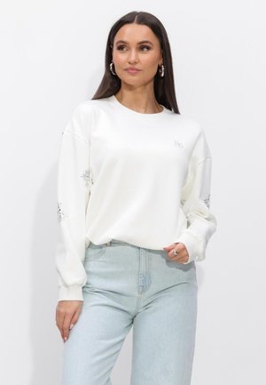 Vrouw met lang donker haar, zilveren oorringen in de vorm van ringen, een witte trui met subtiele mouwdetails en lichtblauwe spijkerbroek, staande tegen een witte achtergrond.