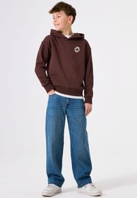 Sudadera con capucha marrón y un logo en la parte frontal, combinada con unos vaqueros azul holgados y zapatillas blancas. Conjunto casual en una persona de pie.