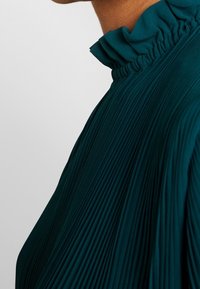 Gros plan d'une personne portant un chemisier plissé bleu sarcelle foncé avec un col haut à volants sur un fond uni.