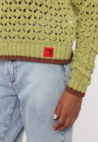 Pull vert tricoté avec bordure marron et étiquette rouge Max & Co., associé à un jean bleu clair, une main reposant le long du corps.