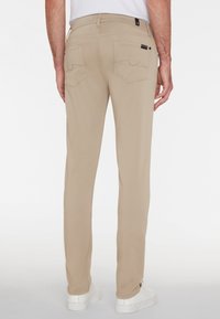 Beige Hosen im Slim-Fit-Stil mit zwei Gesäßtaschen. Mit verstärkten Nähten und einem schwarzen Markenlabel in der Nähe des Bundes. Kombiniert mit weißen Sneakern.