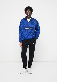 Man in een blauw gestreepte Nike Air pullover, zwarte Nike joggers en witte sneakers, staand tegen een plain witte achtergrond.