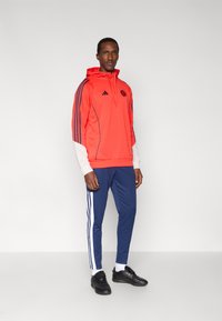 Sweat à capuche rouge de sport avec des rayures et un logo bleu marine, assorti à un pantalon de survêtement bleu marine avec des rayures blanches sur les côtés. Des chaussures de sport noires complètent la tenue.