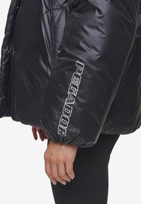 Zwarte pufferjack met gequilte textuur, elastische manchetten en opvallende witte opdruk "PEGADOR" op de mouw. Gladde materiaalafwerking.