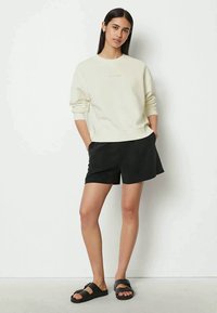 Young woman with long dark hair stands with hands in pockets, wearing a cream sweatshirt, black shorts, and black slide sandals against a white wall.