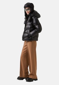 Zwarte pufferjack met een capuchon, met een glanzende textuur en een rits. Gecombineerd met brede bruine broeken en stevige zwarte laarzen.