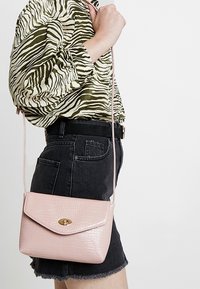 Crocodile-textured pink crossbody bag with a flap and gold hardware, hanging from a long strap, worn against a black denim skirt.