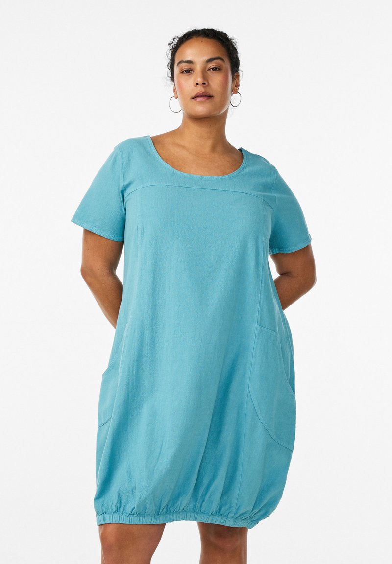 Donna che indossa un vestito azzurro chiaro, corto e a maniche corte con tasche, in piedi contro uno sfondo bianco semplice con le mani dietro la schiena.