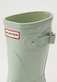 Botte en caoutchouc vert menthe avec un haut arrondi, un bord texturé, une boucle sur le côté et une étiquette de marque sur fond rouge.